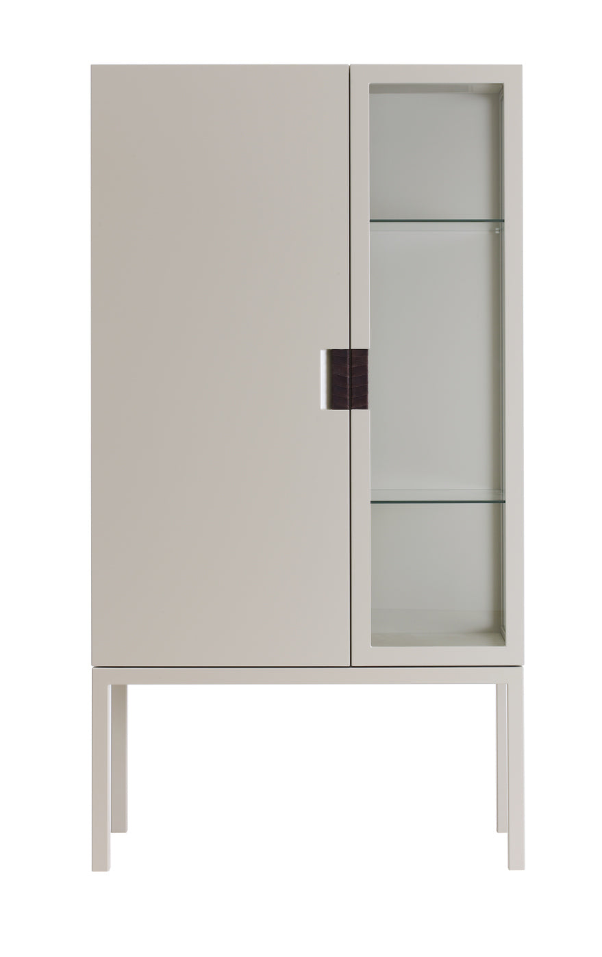 Designed by Anya Sebton and Eva Lilja Löwenhielm 2014.  Glass cabinet in lacquered MDF, stand in lacquered metal. The left side has a covered door and a covered side with two lacquered shelves and the right side has a glass door and a glass side with two glass shelves. Doors have twined leather handles.